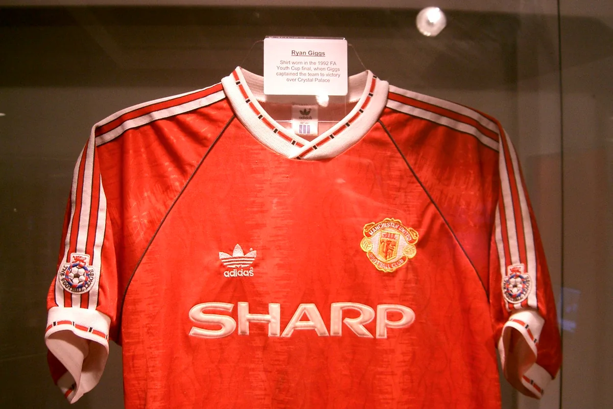 Kostum merah Manchester United dengan logo Adidas dan sponsor SHARP di etalase museum, dengan label Ryan Giggs