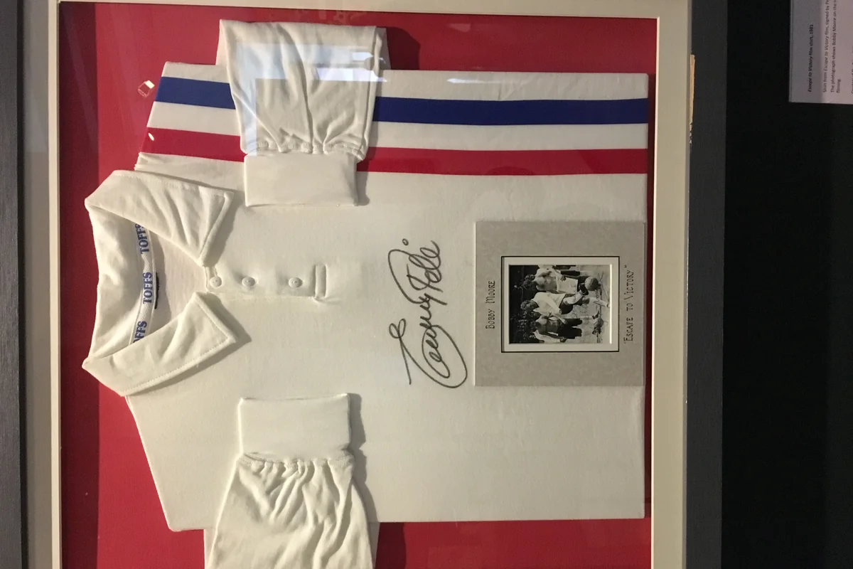 Kostum putih bertanda tangan dengan garis merah biru di sisi, dibingkai di dinding museum