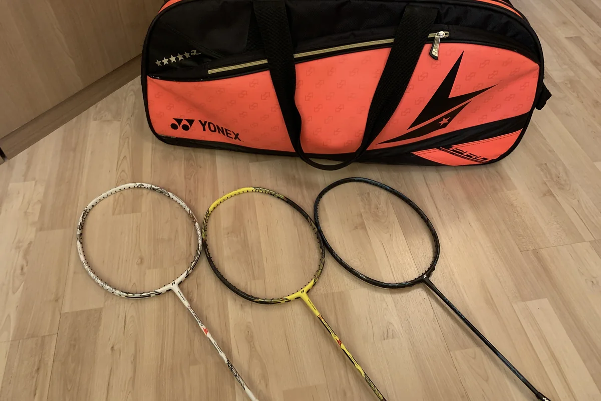 Tiga raket Yonex putih, kuning, dan hitam tergeletak di lantai kayu di depan tas raket Yonex oranye