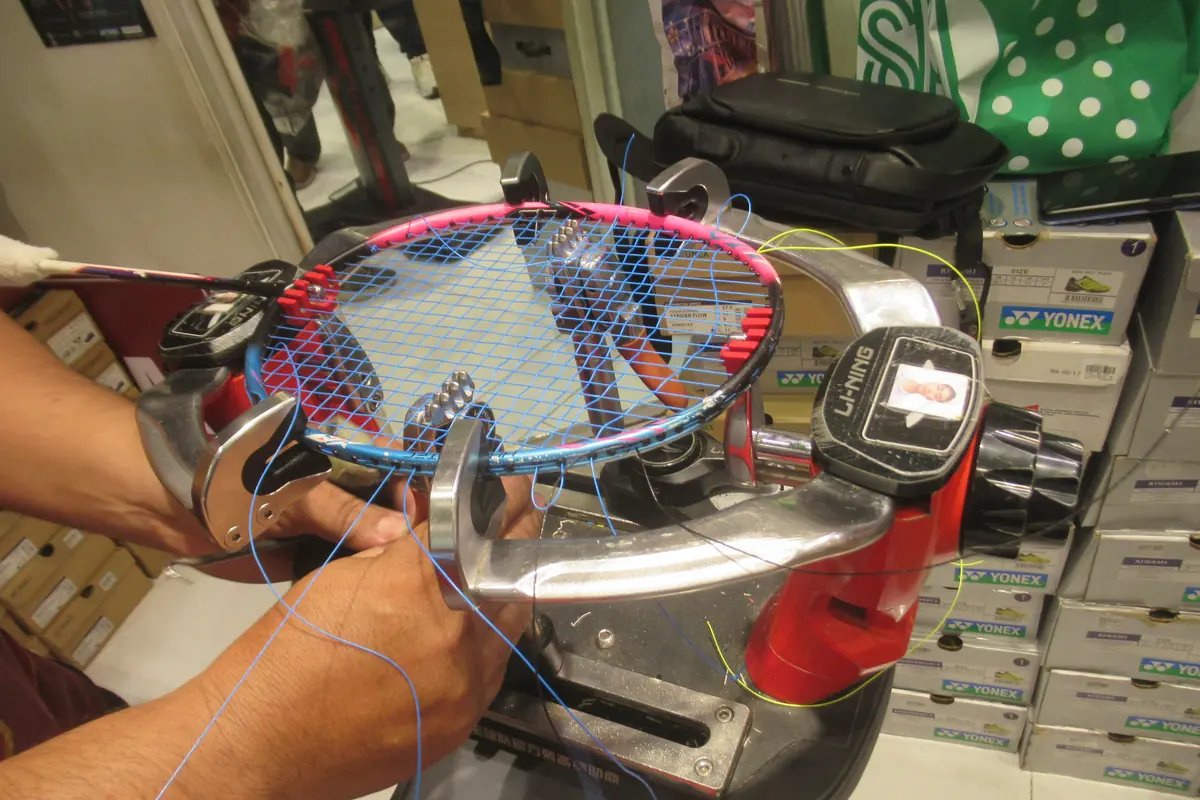 Tangan petugas memasang senar biru pada raket badminton merah muda di mesin pasang senar Li-Ning
