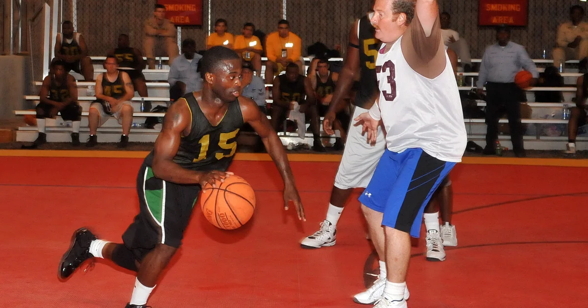 Pemain basket berkaus hijau menggiring bola melewati lawan berkaus putih di lapangan indoor merah