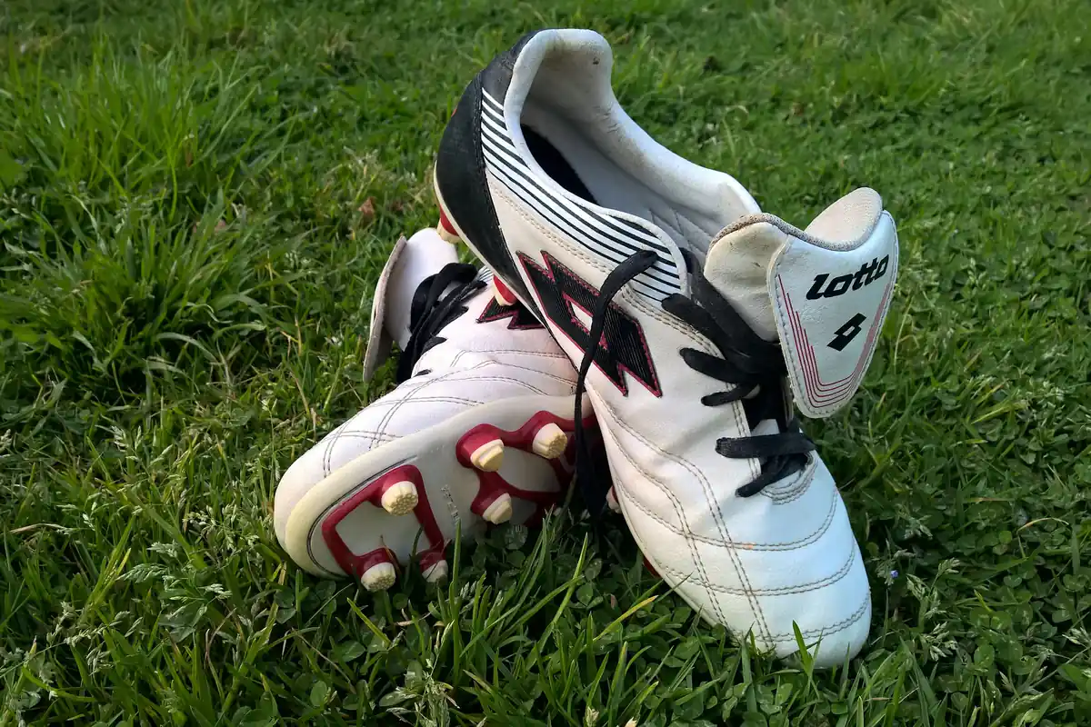 Sepasang sepatu sepak bola Lotto putih hitam dengan pul di bagian bawah, tergeletak di atas rumput