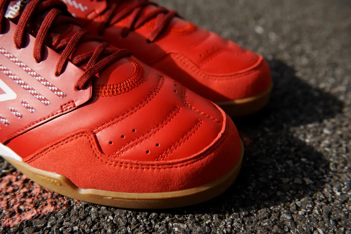 Close-up bagian depan sepatu futsal Umbro merah dengan jahitan pada upper dan lapisan suede di ujung sepatu