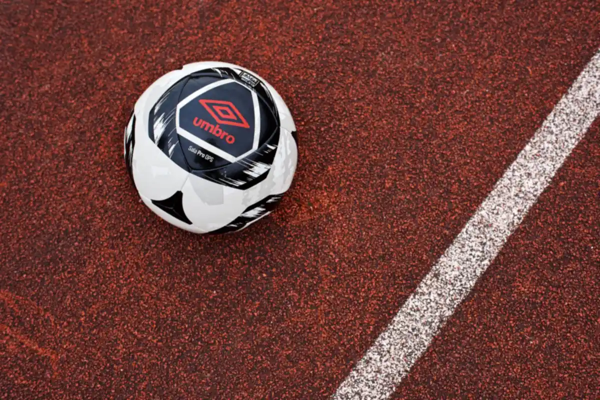 Bola futsal Umbro putih hitam dengan logo Umbro merah di atas lantai lapangan berwarna merah bata bergaris putih