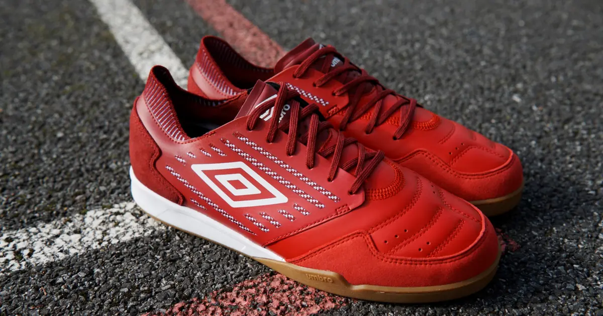 Sepasang sepatu futsal Umbro merah dengan logo berlian Umbro putih dan sol karet cokelat, diletakkan di permukaan lapangan bergaris