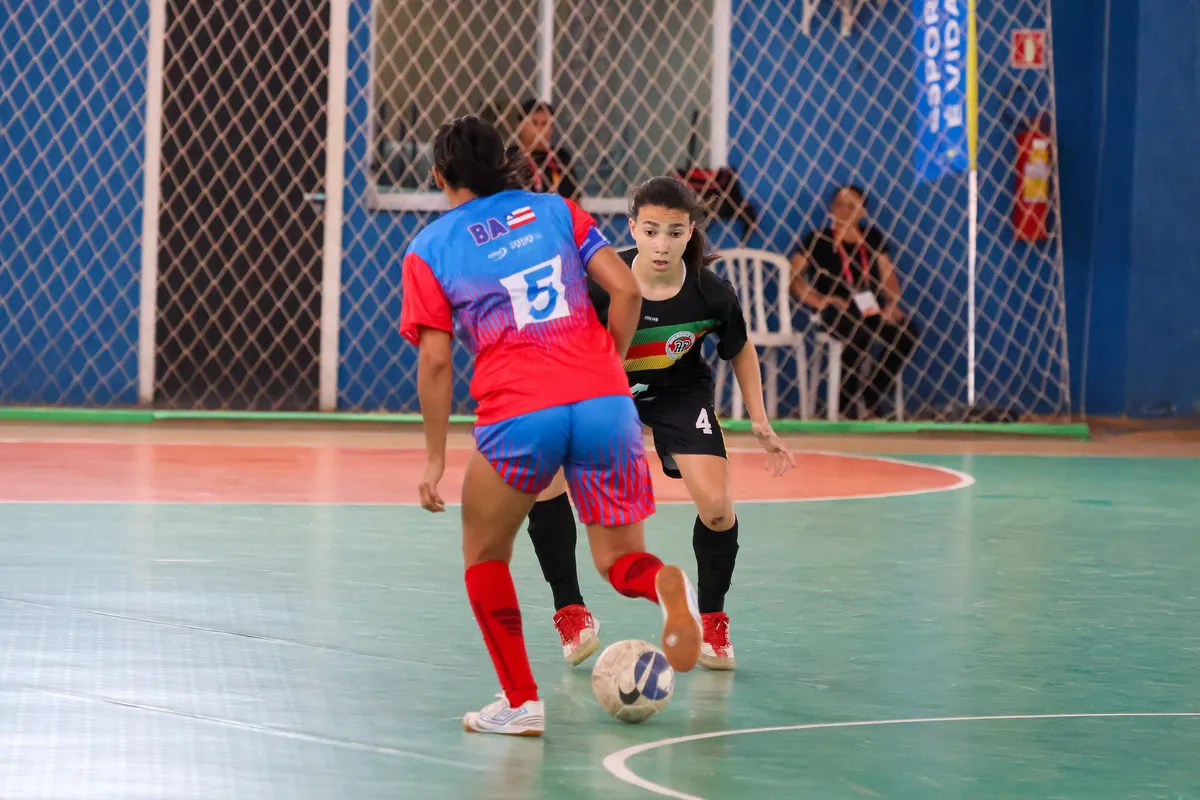 Dua pemain futsal putri berebut bola di lapangan indoor hijau pada Jogos da Juventude 2025 di Brasil