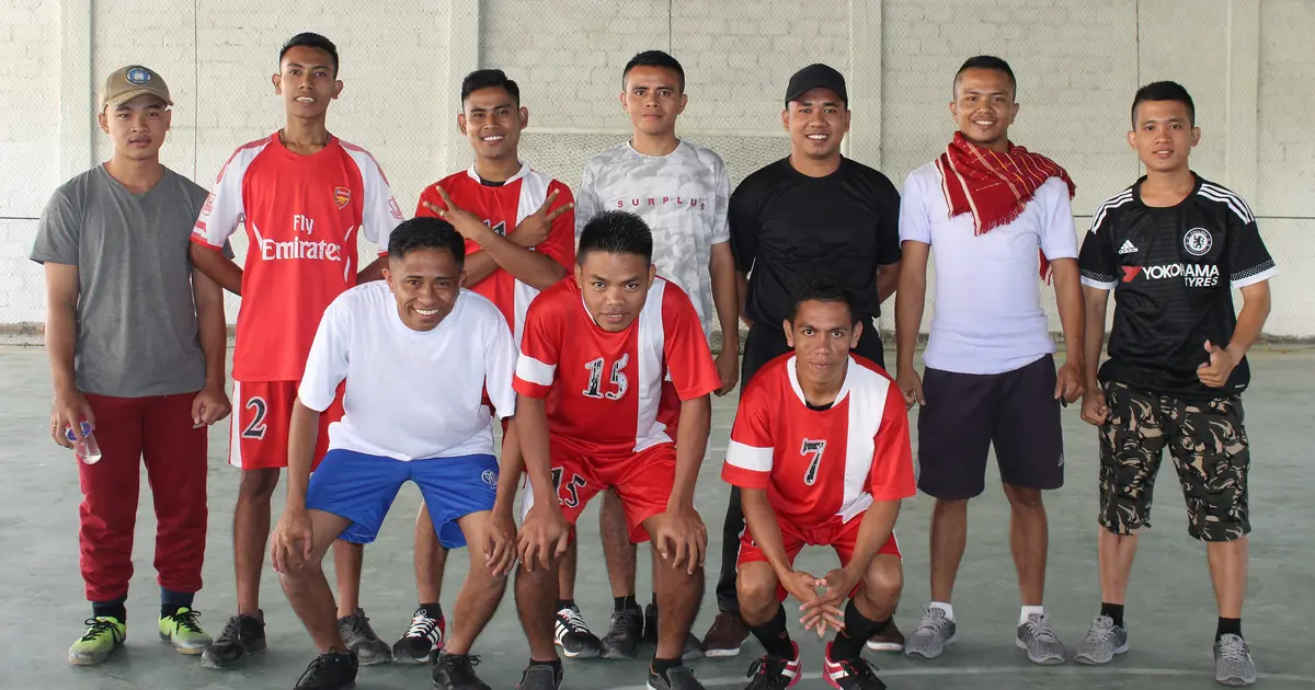 Sepuluh pemuda Indonesia berfoto bersama di lapangan futsal beratap setelah pertandingan, sebagian memakai jersey merah