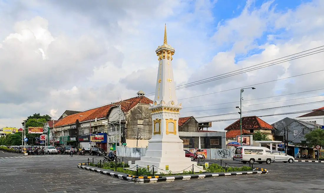 Monumen Tugu Yogyakarta berwarna putih dan emas di tengah persimpangan jalan, dengan deretan ruko dan kendaraan di sekitarnya